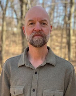 Headshot of man in gray shirt outside with trees in the background.